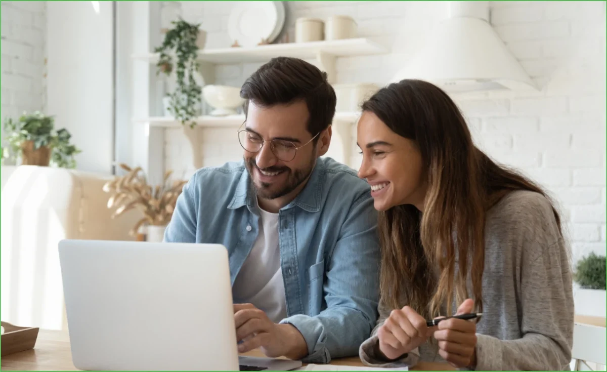 A couple sitting together at a kitchen table, smiling as they read reviews on a laptop.